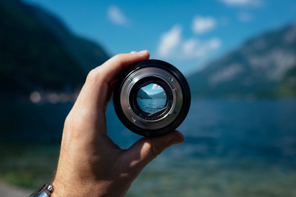 a hand holding a camera lens with a body of water in the background