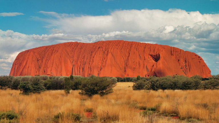 a large red rock in the desert with Uluru in the background