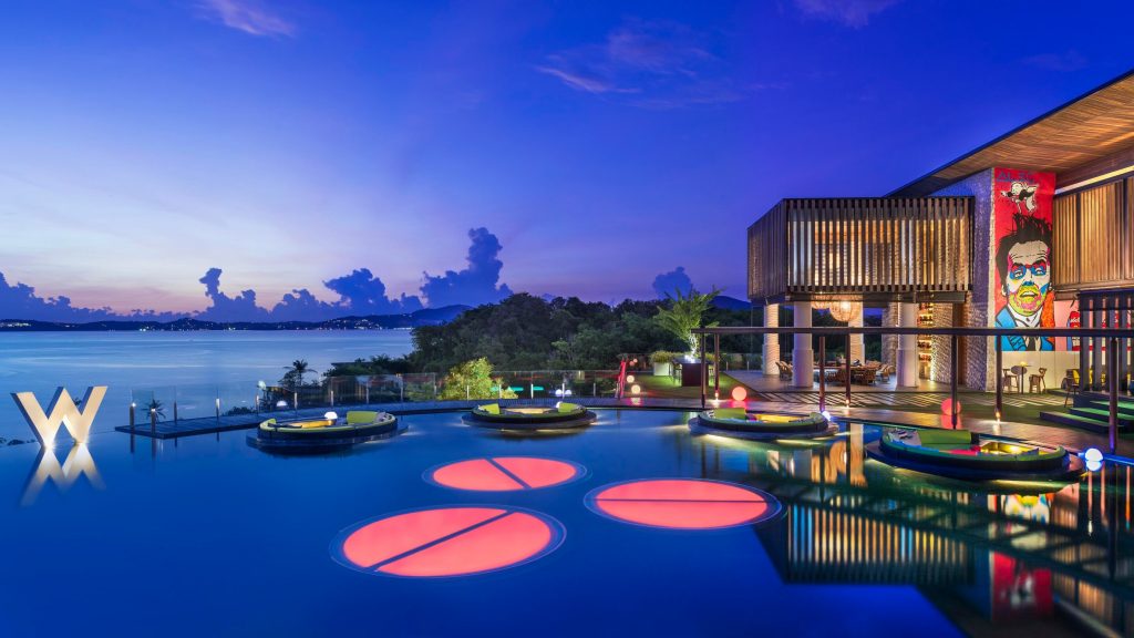 a pool with floating seats and a building with trees and a blue sky